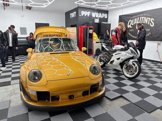 A golden yellow widebody Porsche next to a white sports bike in a detailing bay, with people mingling about.