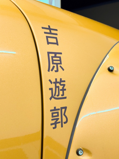 The Kanji for “Yoshiwara Red Light District” (吉原遊郭) written vertically on the fender of a golden yellow car.