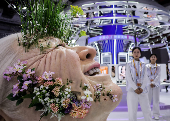 Young women look at the artificial giant head of an elderly woman with flowers and grass growing through skin,