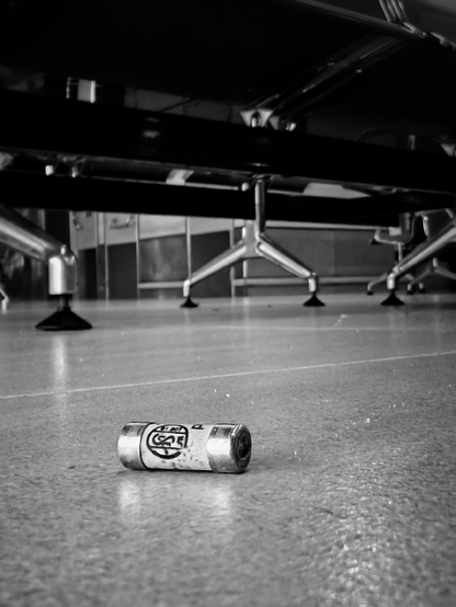 A black and white photo showing an electric fuse lying on the floor beneath metal chairs on a tiled floor.