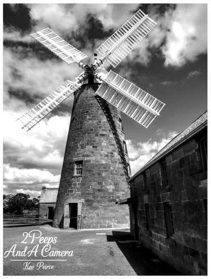 Black and white photograph of the historic Oatlands Windmill in Tasmania.

The tall, circular stone windmill is captured from a low angle, with its large sails angled upwards against a dramatic, cloudy sky.

An adjoining two story stone building is visible on the right, casting a sharp shadow. The ground is gravel and dirt.