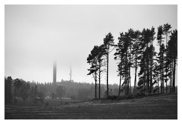 En liggande svartvit bild på ett landskap med en träridå av tallar till höger. På vänster sida längre bort står etthus och längre bort i skogen står en sändarmast och en mobilmast till hälften synliga i dimman och regnet.