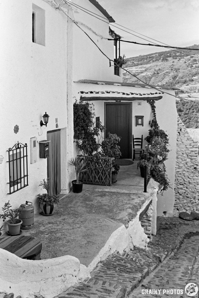 Black and white photo of a whitewashed house in Capileira with potted plants, a chair, and a small patio on a narrow cobbled street, set on a hillside with distant mountains in the background.