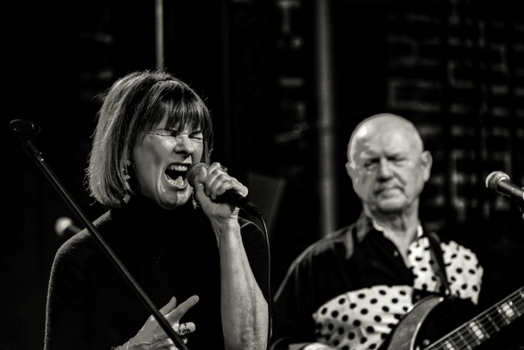 Das Bild zeigt eine Sängerin mit zu einem Schrei aufgerissenen Mund.
Sie hält ihr Mikrofon in der Hand. Rechts im Bild ist etwas unscharf ein Gitarrist zu sehen.