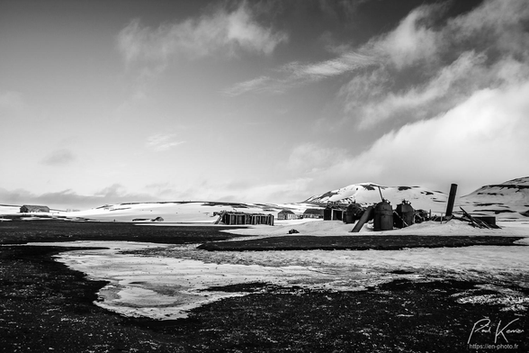 photographie monochrome en noir et blanc de l'ancienne station baleinière sur Deception Island captée depuis le rivage de sable noir encore partiellement recouvert de neige… Parfait pour du noir et blanc.
Quelques nuages occupent le ciel sur la droite, au-dessus des ruines de l'ancienne station avec notamment des réservoirs cylindriques métalliques, une cheminée également en métal, plus loin, des bâtiments en bois et avec en arrière-plan des collines encore enneigées.