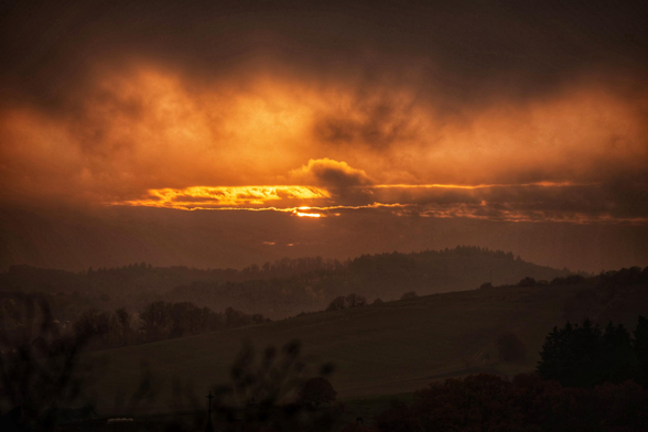 Das Bild zeigt einen Sonnenuntergang über einer hügeligen, bewaldeten Landschaft.
Die Sonne ist als leuchtender Streifen sichtbar, der durch eine Wolkenschicht bricht.
Der Himmel ist in intensive Orange- und Rottöne getaucht, während die Wolken dunkel und dramatisch wirken.
Die Silhouette der Hügel und Bäume ist im Vordergrund zu erkennen.