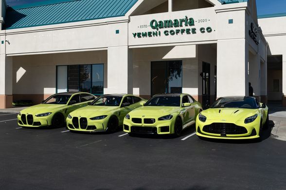 Four sports sedans parked outside a shop called “Qamaria Yemeni Coffee Co.” The cars are all in a greenish-yellow color, but the Aston’s color is ever so slightly more yellow.