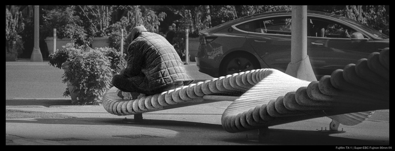A person sits hunched over on the end of a bench built in a wavy, rippling motion.  They are holding an ebook reader in their hands and looking down at it.  The surroundings are generally empty save for a car passing by in the background.