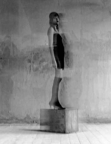 A black and white photo of a woman in a short dress and high heels stands atop a wooden block.
