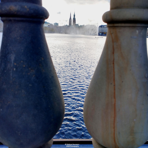 View over lake Binnenalster in Hamburg, Germany, towards two steeples on the Eastern shore seen through two columns of the stone trellis on Lombardsbrücke.

© Stefanie Neumann - #KBFPhotography