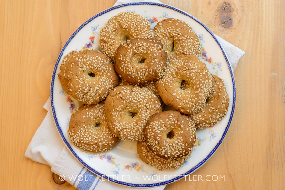 The photograph shows a plate of Greek olive oil cookies. They are shaped like rings, round with a hole in the middle, and covered in sesame seeds.