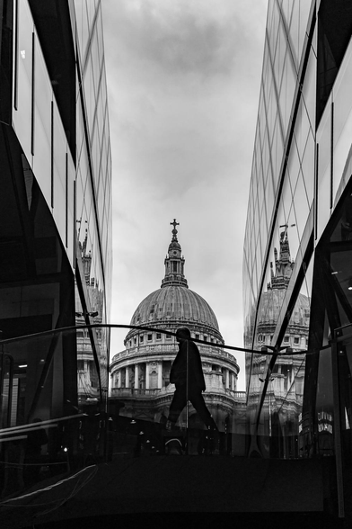 The image is a black and white photograph showcasing a juxtaposition of modern and historical architecture. The composition is symmetrical, with two modern glass-clad buildings flanking the view. These buildings are tall and sleek, their surfaces reflecting the surrounding environment, including the sky and the iconic St. Paul's Cathedral.

A silhouetted figure of a person is walking in front of the cathedral, adding a human element to the otherwise architectural focus. The person's form is simple, and the lack of detail draws attention to the overall composition and the play of light and shadow.