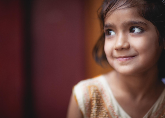 A young girl in Chandni Chowk, Old Delhi, glances sideways with a shy smile, her sequinned dress catching soft afternoon light against a deep red and ochre background.