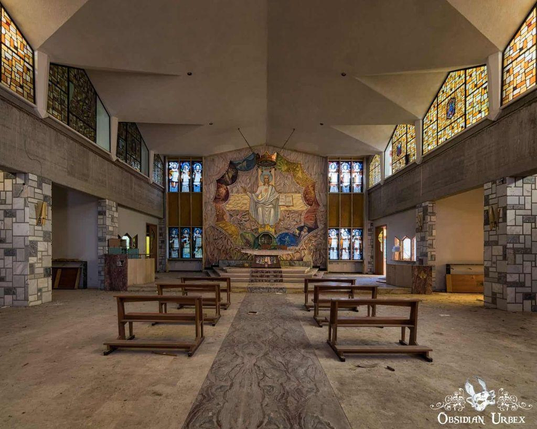 An empty church interior with wooden pews, stained glass windows, and a large colorful mural behind the altar.