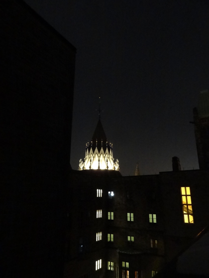 A night time picture of the brightly lit spire at the top of the Library of Parliament from within a conclave of dark buildings, some with dimly lit windows.