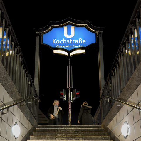 Motiv:
Das Foto zeigt den Eingang zur Berliner U-Bahn-Station Kochstraße, die auch den Zusatz „Checkpoint Charlie“ trägt. Im Vordergrund sind Treppenaufgänge zu sehen, die von zwei Personen genutzt werden. Über dem Treppenaufgang hängt das beleuchtete Stationsschild in Blau und Weiß.

Stimmung & Wirkung:
Die Aufnahme vermittelt eine urbane, nächtliche Atmosphäre. Das Licht der U-Bahn-Beleuchtung kontrastiert mit der Dunkelheit des Himmels. Die Szene wirkt ruhig und geordnet, mit einem leichten Gefühl von Bewegung durch die gehenden Personen.

Technische Details:
Farbe: Farbaufnahme mit dominanten Blautönen des Schildes, neutralen Grautönen der Architektur und warmem Kunstlicht.
Schärfentiefe: Große Schärfentiefe, wodurch Schild, Treppe und Personen gleichermaßen deutlich erkennbar sind.
Lichtverhältnisse: Künstliche Beleuchtung bei Nacht, gleichmäßige Ausleuchtung durch Lampen und das beleuchtete U-Bahn-Schild.