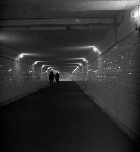 The black-and-white photo shows a gloomy pedestrian tunnel under the railway tracks, stretching into the distance. The tunnel is tiled and illuminated by small light bulbs with grates at the top of the side walls. In the distance, a bright exit can be seen, with two dark figures in the background.