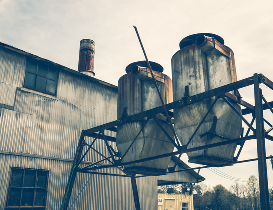 An industrial structure featuring two weathered cylindrical metal silos mounted on an angular steel framework against an overcast sky. The silos show significant rust and corrosion, with reddish-brown staining on their silver-gray surfaces. Each silo has a domed top with protruding pipe fittings. The supporting framework consists of dark metal beams arranged in a triangular configuration. To the left stands a corrugated metal building with vertical ribbing and a multi-paned window. A thin vertical rod extends upward from the central silo. The scene has a muted, desaturated color palette. Bare winter trees are visible in the background along with what appears to be a white shipping container or storage unit.