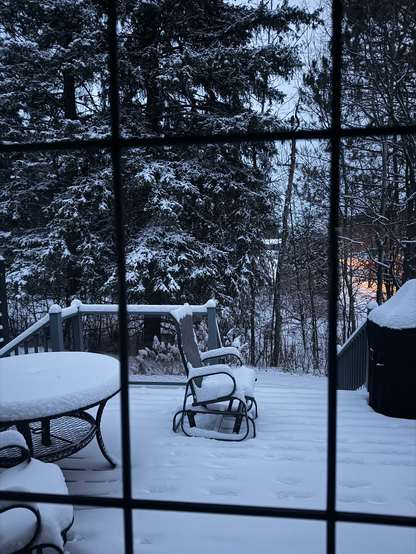 Snow covered patio. Table, rocking chair and railing with snow covered trees in backyard. Sun rising over the river in background .