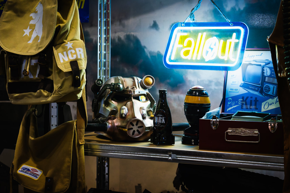 In a dimly lit room, a collection of items related to the video game “Fallout” is displayed. A yellow backpack with “NCR” and a bear logo hangs on a metal shelf. Below it, a detailed replica of a power armor helmet rests next to a dark cola bottle. To the right, there’s a round object resembling a miniature atomic bomb. A blue neon sign glows with the word “Fallout,” casting colorful light over a Pip-Boy kit box and a metallic case.