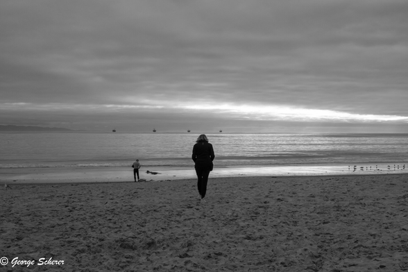 Black and white image of sunset at Carpinteria State Beach on a cloudy day .  The foreground is a sandy beach.  In the background is the ocean, and on the horizon the Channel Islands are visible, along with a row of oil platforms.  In the center of the image a woman, seen from behind in silhouette, is walking towards the water.