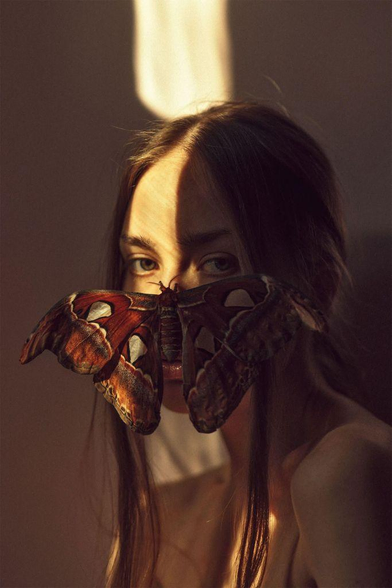 A person with long hair is adorned with a colorful butterfly resting gently on their face, illuminated by soft, warm light.