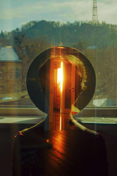 A person's silhouette reflects a doorway with bright sunlight, framed by a window, creating a surreal, introspective scene.