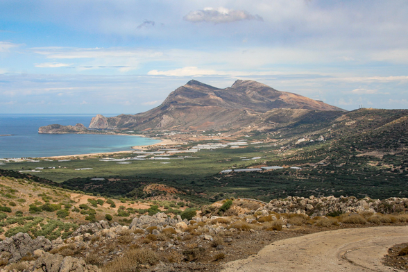A panoramic view of a coastal landscape featuring mountains, a bay with turquoise water, and lush greenery in the foreground. The sky is partly cloudy, adding to the scenery's natural beauty.