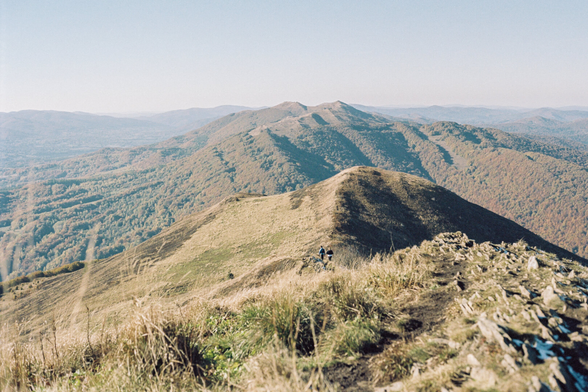 A couple walking along a trail through hills covered with dry grass and rocks, with a sunny sky in the background