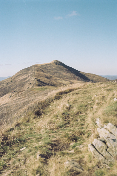 Mountain hills covered with dry grass, grass and rocks in the foreground, and a trail leading to the summit in the background.