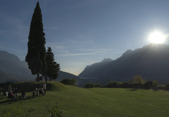 Landscape photograph of a lawn overlooking a lakeside town between mountains, taken during daytime. The sun is glistening just above the top of one of the summits bordering the town with its rays projecting through the contours of the mountain. On the lawn at the left side of the photograph, a few people are sitting on garden chairs by tables from an outdoors cafe. Behind these people, a tall pine tree dominates the picture, with at its base a bench on which two people are sitting close together, facing the landscape view.