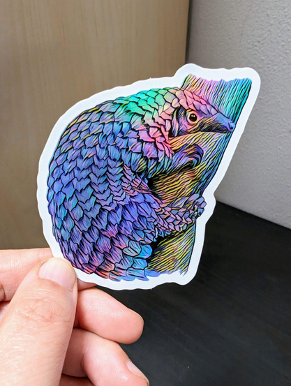 Photo of a left hand holding a diecut sticker. The sticker is a holographic pangolin drawing.