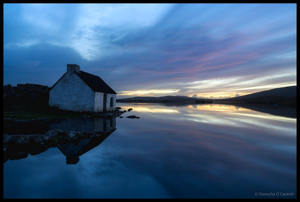 An abandoned white-walled fishing hut with dark roof beside perfectly still water reflecting a dramatic twilight sky with pink and blue clouds at Screebe in Connemara, County Galway, with silhouetted mountains visible on the horizon.