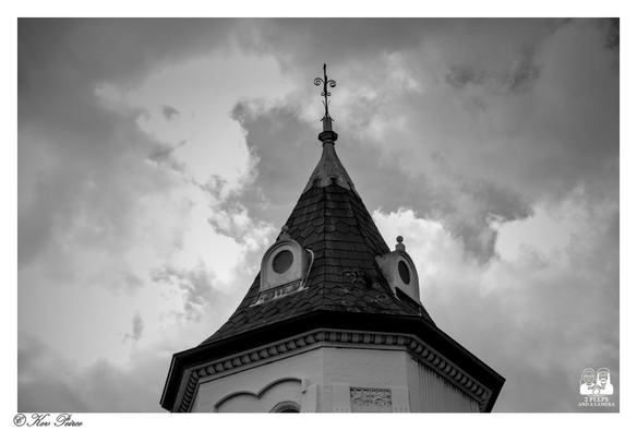 A dramatic, high contrast black and white photograph of the turret and spire of a historic building in Launceston, Tasmania, set against a brooding, cloudy sky.

The octagonal turret has a conical, tiled roof, small circular windows, and is topped with a decorative metal finial or weather vane against the textured clouds.