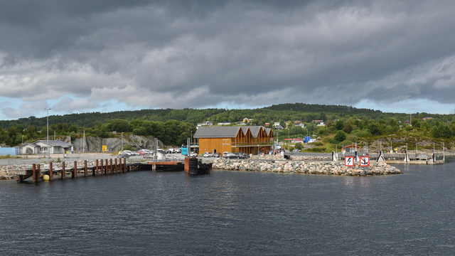 A photo of a ferry terminal with a wood clad main building. There a green hills in the distance. The sky is filled with grey clouds.