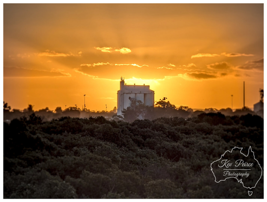 A wide angle landscape photo taken at sunset. The sky is a gradient of soft orange and yellow, silhouetting the horizon.

In the middle distance, a bright white grain silo and other industrial structures stand above a thick band of dark green and grey-brown Australian native scrub and low-lying vegetation in the foreground.