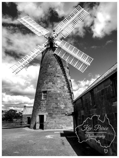 Black and white photograph of the historic Oatlands Windmill in Tasmania.

The tall, circular stone windmill is captured from a low angle, with its large sails angled upwards against a dramatic, cloudy sky.

An adjoining two story stone building is visible on the right, casting a sharp shadow. The ground is gravel and dirt.