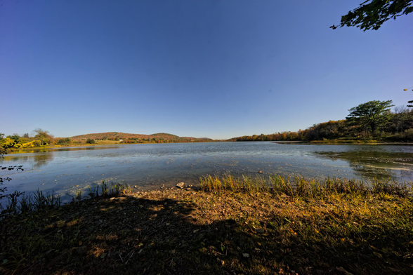 The horizon is just below the centerline where a lake disappears into a line of hills under a huge empty blue sky reflected in a quiet lake with a stone shore