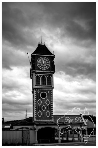 A tall, four sided clock tower in Esperance, rendered in black and white.

The tower features a brick base with diamond patterns, arched windows, and a clock face near the top, all under a dark, dramatically cloudy sky.

The image is signed 'Kev Peirce'.