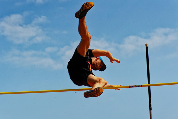 A man is frozen in air with hisfeet facing us and his hands still behind the yellow horizontal bar,  a green pole that got him there is resting on the far side and there are thin clouds in the far distance