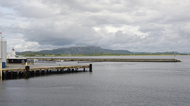 A photo of a pier and breakwater with low rocky mountains in the distance. The sky is filled with clouds.