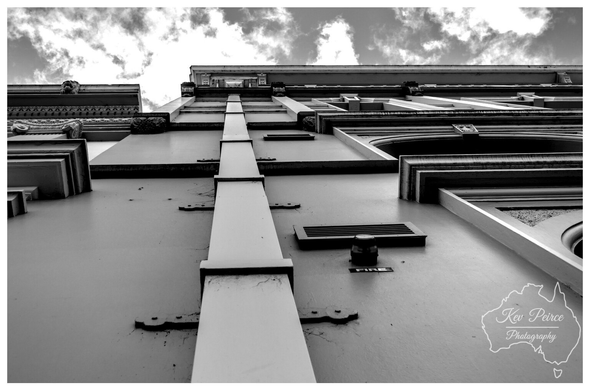 Black and white, low angle photograph of the facade of a tall, classic building.

A vertical downpipe runs up the center, emphasizing the height and leading the eye toward the dramatic, cloudy sky. 

Signed by Kev Peirce Photography.
