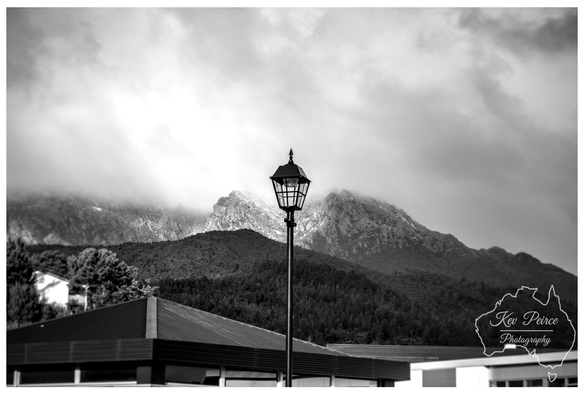 A moody black and white photograph featuring a classic street lamp centered against a dramatic mountain landscape, in Queenstown.

The foreground shows the roofline of a modern building. Behind the light, a forested slope rises to meet rugged, rocky peaks shrouded in low-hanging, textured clouds.

The overall composition creates a strong contrast between the man-made structure and the raw natural environment.