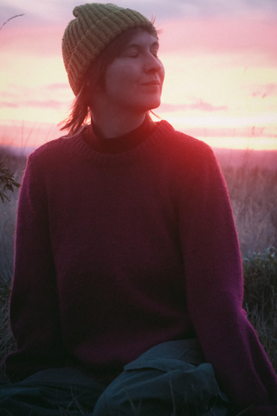 person sitting on a field of grass with their head turned to their left, eyes closed with a red sunset behind them