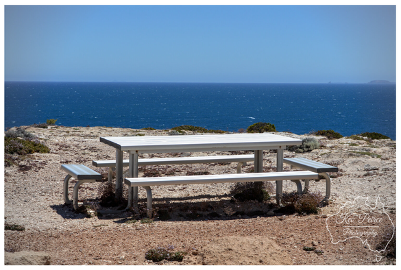 A white metal picnic table and two benches sit on a reddish brown, rugged cliff overlooking a deep blue ocean that meets a bright clear sky.