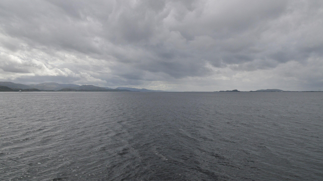 A photo of a wide fjord with dark water and a cloudy sky.