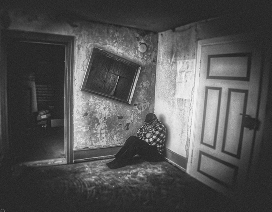 Ein Mann sitzt zusammengekauert in der Ecke von einem alten Zimmer. Schräg über ihn ist ein alter Bilderrahmen an der Wand zu sehen. Bildsprache geht dahin, dass der Mann durch die Depression aus den gewohnten Rahmenbedingungen rausgefallen ist und er es nicht schafft wieder zurück zu gehen.