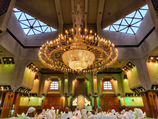 A breathtaking shot from inside the Grand Mosque in Mecca, Saudi Arabia, looking up at the magnificent main chandelier. The grand, circular chandelier, adorned with countless lights, hangs prominently from the ceiling, illuminating the vast prayer hall with a warm, golden glow. Below, numerous worshippers dressed in white are seated on the carpeted floor, engaged in prayer and reflection. The mosque's architecture features intricate geometric patterns, arched windows, and high ceilings, creating a serene and spiritual atmosphere. The light from the chandelier is reflected on the shiny marble floors and wooden accents, adding to the grandeur of the space.