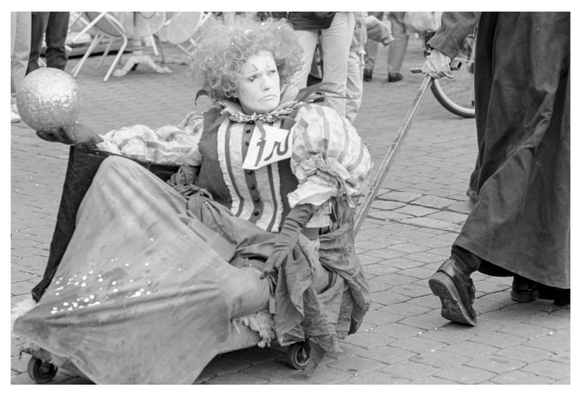 Schwarzweißfoto im Querformat. Eine Szene aus einem Straßenthater. Ein Schuauspieler sitzt bzw. liegt auf einem Karren. In einer Hand hält eine Kugel (königliches Symbol) und auch die Kleidung ist eher herrschaftlich. Die Perrücke ist aber sehr wild und die Schmicke passt eher zu einem Narr.