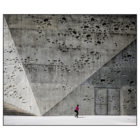 A person wearing a bright pink or magenta jacket and dark pants walking across a large concrete plaza. The dominant feature is a massive brutalist concrete wall that fills most of the frame, characterized by hundreds of small rectangular perforations or openings scattered across its surface in an irregular pattern. The wall has geometric angles and planes, with a diagonal line cutting across the composition. On the left side, there are concrete stairs leading upward. A door is visible in the lower right portion of the wall. The stark contrast between the small human figure and the imposing architectural structure emphasizes the monumental scale of the building. (draft by claude.ai)